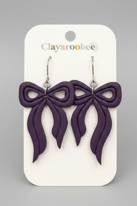 A pair of purple bow-shaped dangle earrings on a white Clayaroobee display card
