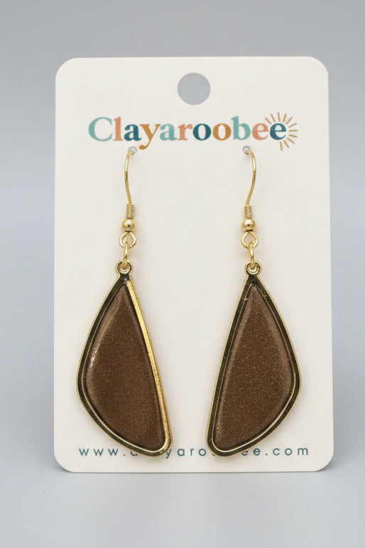 Teardrop-shaped brown clay earrings with gold borders and gold fishhook wires, displayed on a white Clayaroobee card, set against a dark gray wood background.
