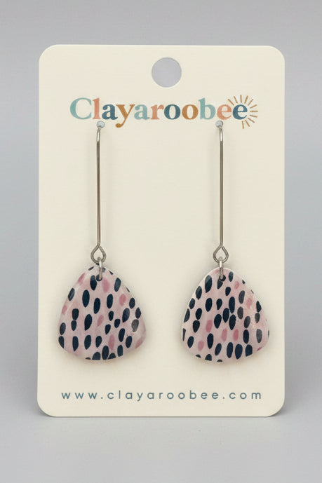 A pair of teardrop-shaped clay earrings with a pink, white, and navy spotted pattern, displayed on a white Clayaroobee brand card against a dark gray wooden surface.