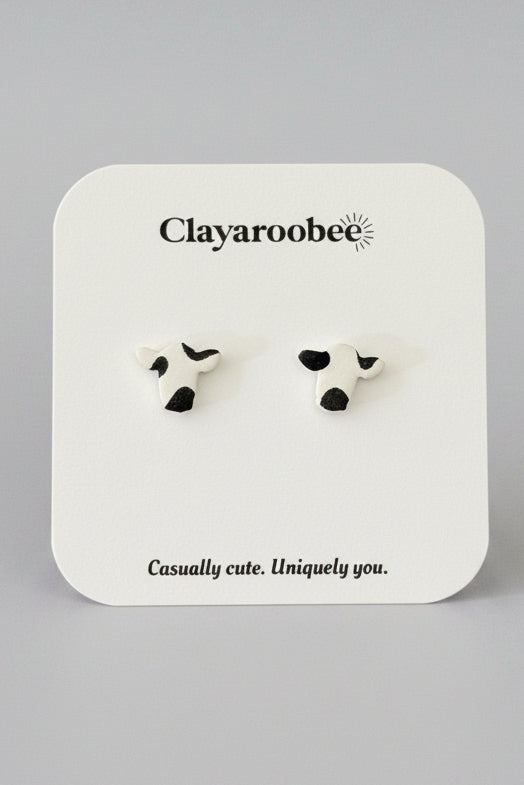 A pair of cow-print stud earrings on a white display card with the brand name Clayaroobee