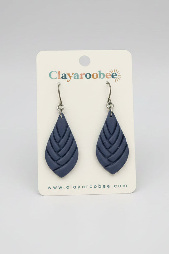 A pair of teardrop-shaped clay earrings in a dusty navy blue color, displayed on a white brand card for Clayaroobee. 