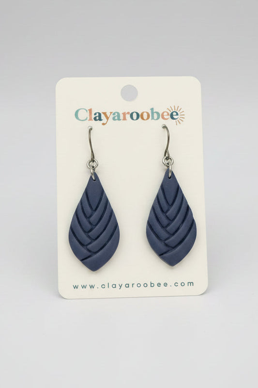 A pair of teardrop-shaped clay earrings in a dusty navy blue color, displayed on a white brand card for Clayaroobee. 