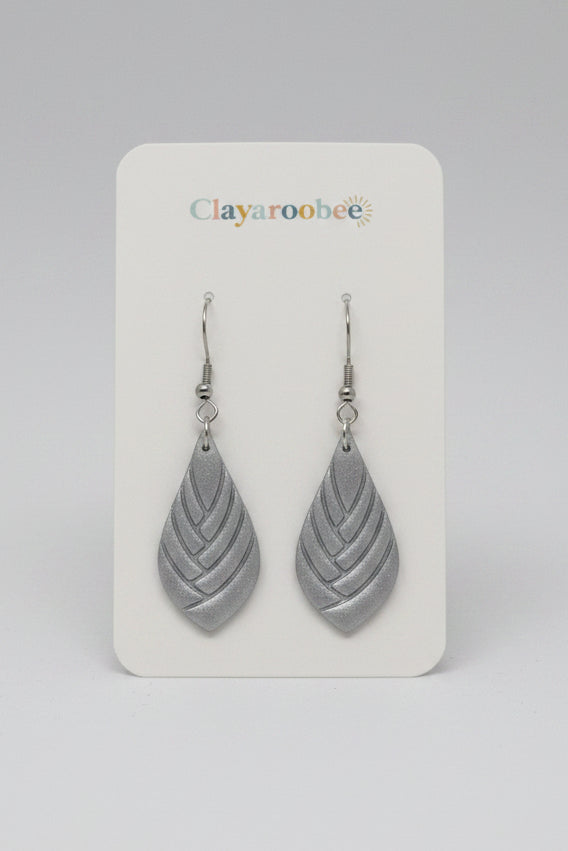 A pair of silver-toned teardrop dangle earrings with a layered leaf-like cutout design, displayed on a white Clayaroobee brand card. 