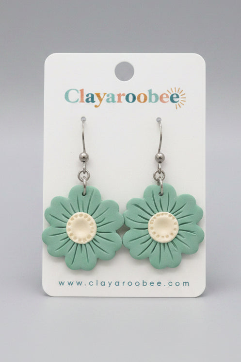 A pair of flower-shaped dangle earrings in mint green with cream centers, displayed on a white clayoobee card on a dark wooden surface.