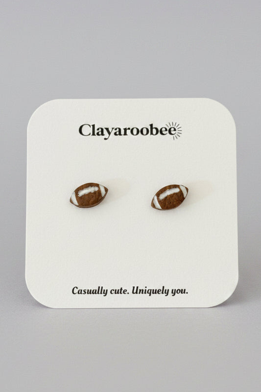 A pair of football-shaped stud earrings on a white display card with the brand name Clayaroobee.