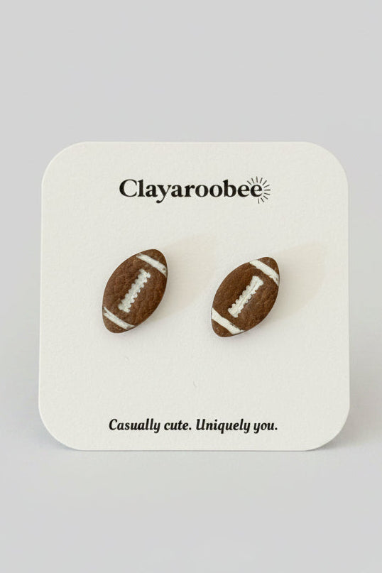 A pair of football-shaped stud earrings displayed on a white branded card. The card shows the brand name Clayaroobee and the text 'Casually cute.