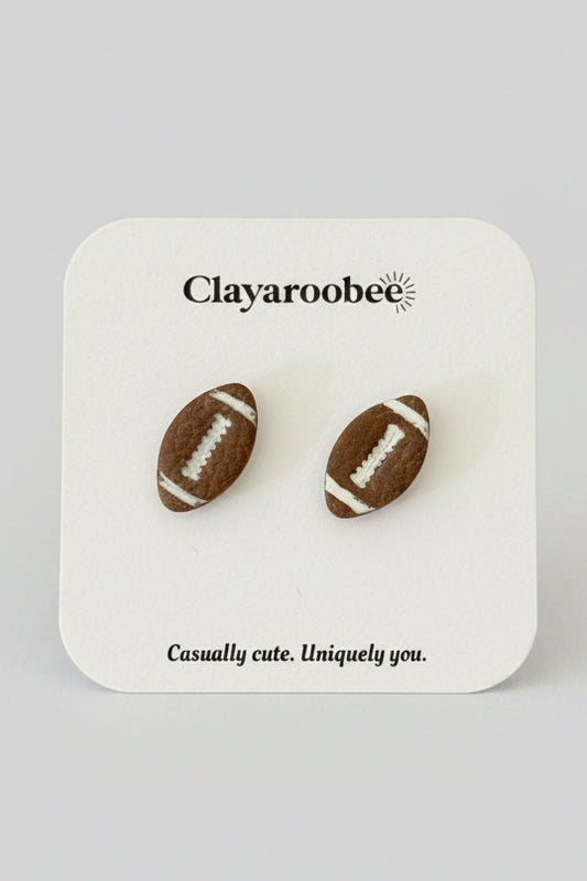 A pair of football-shaped stud earrings displayed on a white branded card. The card shows the brand name Clayaroobee and the text 'Casually cute.