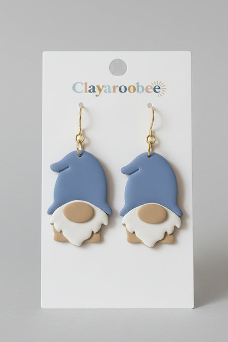 A pair of gnome-shaped drop earrings on a white display card. The earrings feature light blue hats, white beards, and tan faces, hanging from gold hooks.