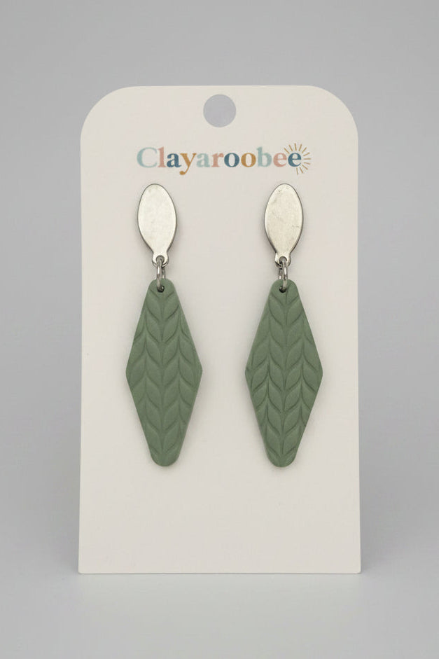 A pair of clay earrings with a sage green textured geometric drop design and silver-tone oval studs, displayed on a white 'Clayaroobee' branded card. 
