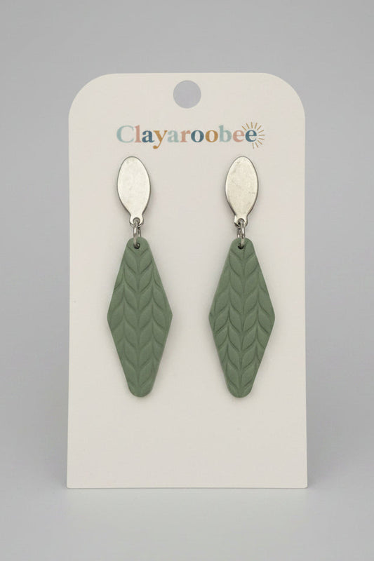 A pair of clay earrings with a sage green textured geometric drop design and silver-tone oval studs, displayed on a white 'Clayaroobee' branded card. 