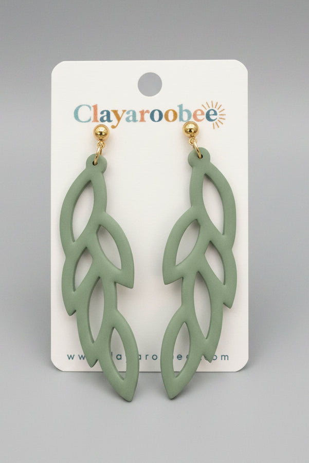 A pair of leaf-shaped dangling earrings in a muted sage green color with gold-tone posts, displayed on a white Clayaroobee brand cardq