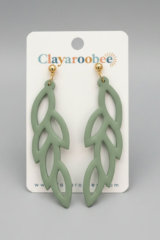 A pair of leaf-shaped dangling earrings in a muted sage green color with gold-tone posts, displayed on a white Clayaroobee brand cardq