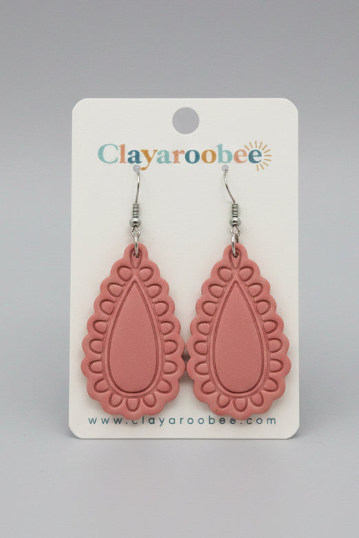 A pair of pink teardrop-shaped dangle earrings with scalloped edges and silver hooks, displayed on a white Clayaroobee brand card. 