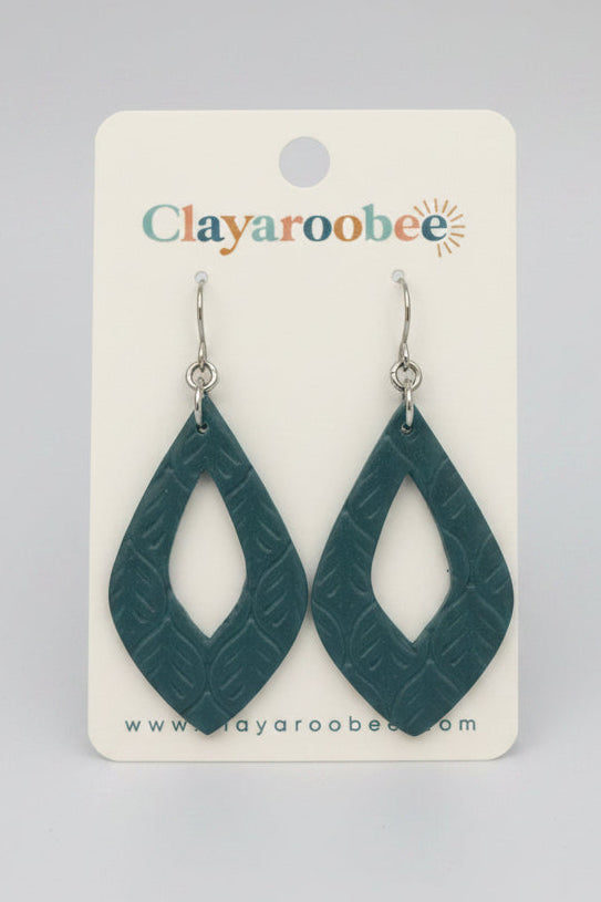 A pair of teal clay dangle earrings with a carved geometric pattern, displayed on a white card labeled 'Clayaroobee' =