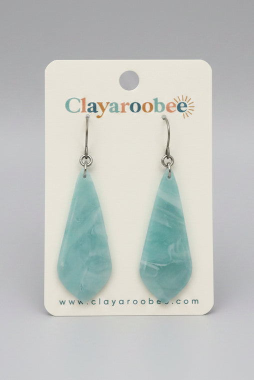 A pair of teardrop-shaped light blue marbled resin dangle earrings on a white Clayaroobee brand card, displayed on a dark gray wooden surface.