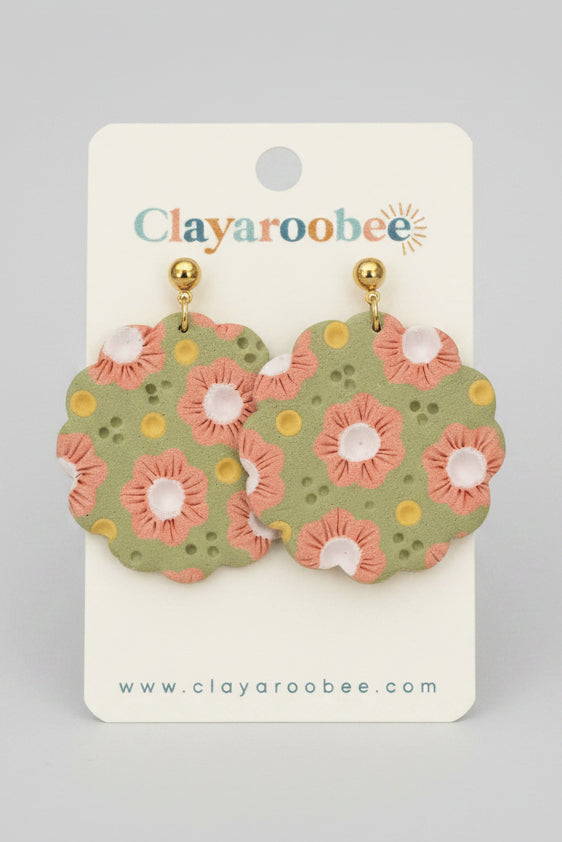 A pair of clay earrings with a green base, pink and white flowers, and yellow dots, hanging from gold posts on a white display card branded Clayaroobee.