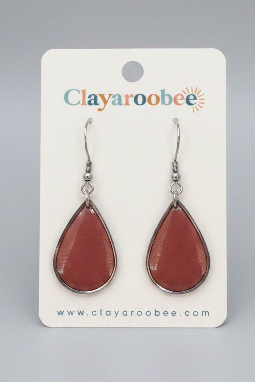 A pair of clay droplet earrings with a terracotta reddish-brown finish and silver hooks, displayed on a white Clayaroobee card. 